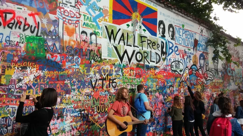 Prag'ın mistik güzelliğini sunan bu seyahat rehberi kapsamında öne çıkan Lennon Duvarı, rengarenk grafitiler, yazılar ve sanat eserleriyle kaplıyken, önünde gitar çalan bir adam ve duvarı fotoğraflayan ziyaretçiler görülüyor.