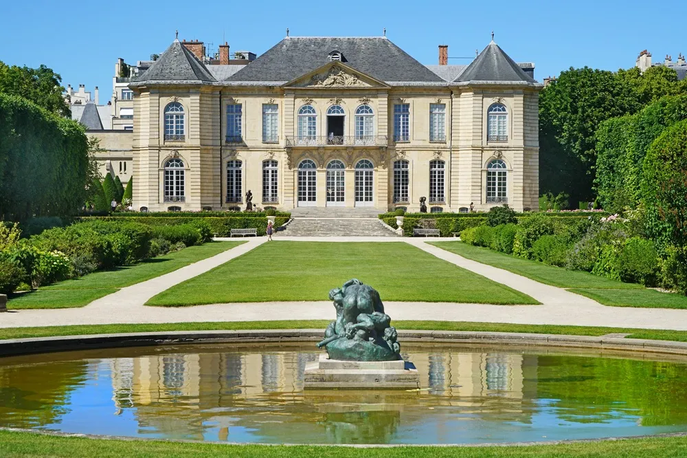 Paris'teki Rodin Müzesi'ne ev sahipliği yapan görkemli Hôtel Biron, önündeki simetrik yeşil bahçesi, düzenli çalıları, ağaçları ve ortasında bronz bir heykelin bulunduğu, binanın ve berrak mavi gökyüzünün yansıdığı dairesel yansıma havuzuyla güneşli bir günde göz alıcı görünüyor.