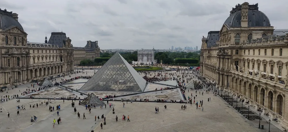Paris'teki dünyaca ünlü Louvre Müzesi'nin geniş, gri bulutlu bir günde görülen avlusunda, merkezi cam piramit ve etrafındaki küçük piramitlerin yanı sıra klasik mimarili görkemli binalar arasında dolaşan yoğun bir kalabalık, uzakta Carrousel Zafer Takı ile şehrin siluetini gözler önüne seriyor.
