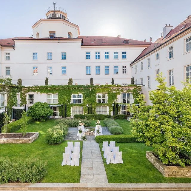 Prag'ın mistik güzelliğini yansıtan, 14. yüzyıldan kalma bir manastırdan dönüştürülen Mandarin Oriental otelinin yemyeşil sarmaşıklarla kaplı beyaz cephesinin önünde, özel bir etkinlik için beyaz sandalyeler ve çiçekli süslemelerle düzenlenmiş sakin ve huzurlu bir bahçe.