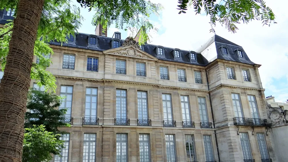 Paris'teki Picasso Müzesi'nin detaylı görünümünde, sol önde dokulu bir ağaç gövdesi ve yeşil yapraklarla çerçevelenmiş, süslü alınlık ve ferforje balkonlu uzun pencereleriyle klasik taş bina ve mansard çatısı net bir şekilde görülüyor.