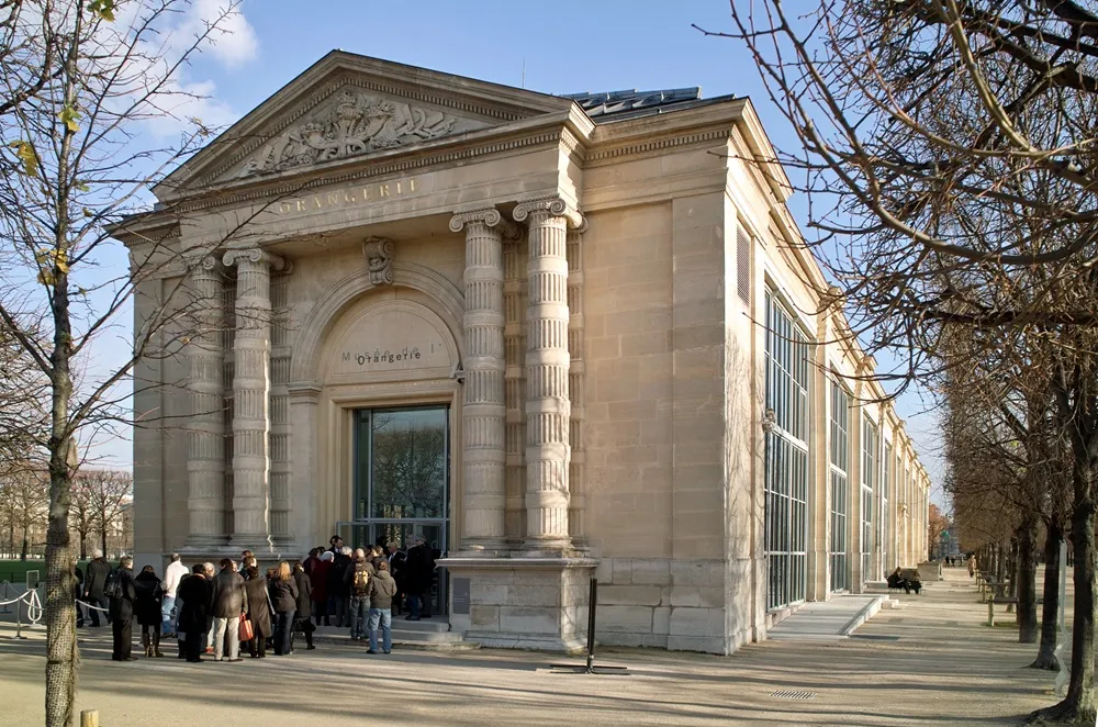 Musée de l'Orangerie yazılı, Neoklasik üslupta, kabartmalı üçgen alınlıklı ve İyonik sütunlarla çevrili kemerli ana girişiyle dikkat çeken bej renkli müze binasının önünde kalabalık bir ziyaretçi grubu toplanmış; yapının sağ tarafında ise boydan boya uzanan pencereler ve mevsimlik dalları çıplak ağaçların dizildiği güneşli bir yol görülüyor.