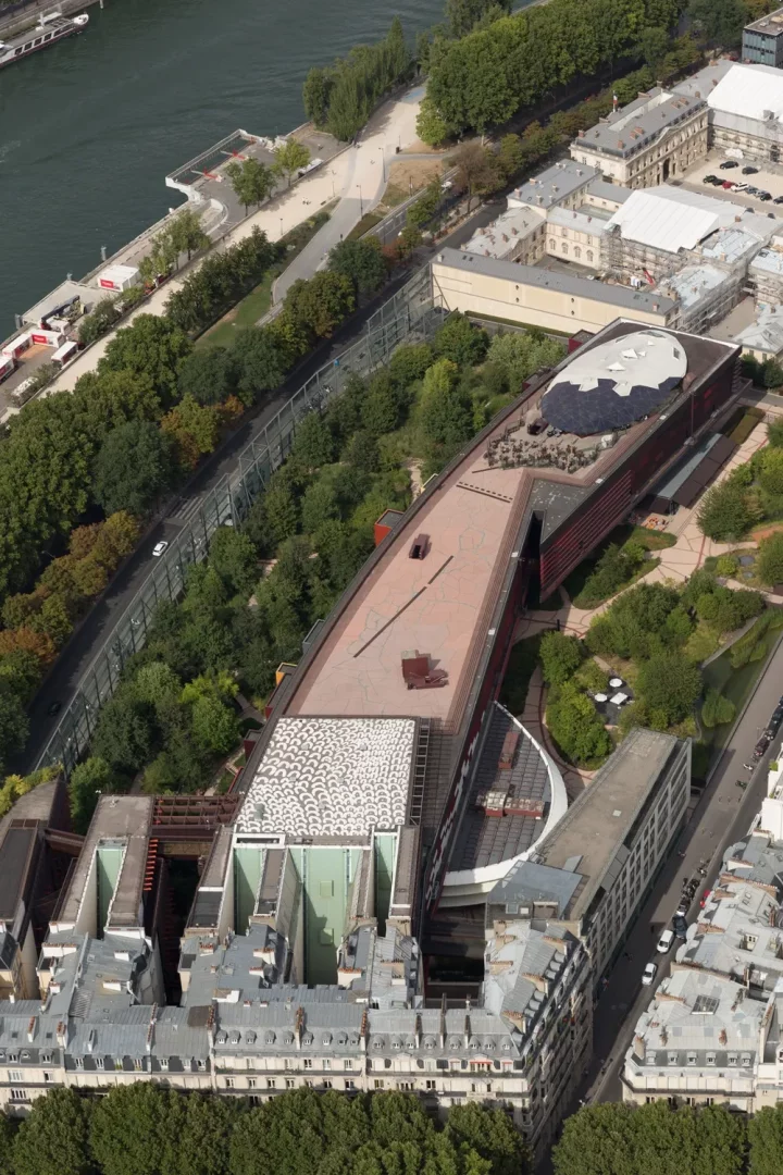 Quai Branly Müzesi'nin, Seine Nehri kıyısında, yemyeşil ağaçlar ve parklarla çevrili uzun, modern mimarili binasının, üzerinde beyaz desenli ve koyu renkli oval çatılar barındıran kompleks yapısının kuşbakışı görünümü.