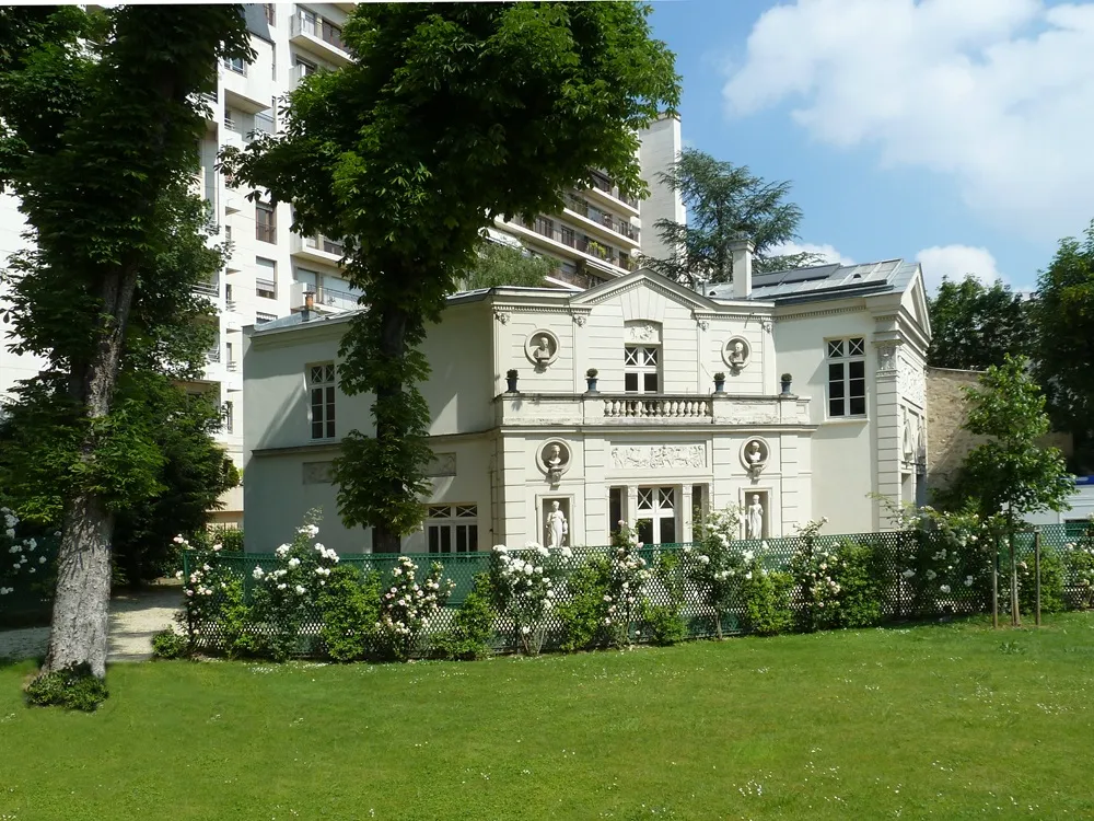 Paris'teki Marmottan Monet Müzesi'nin yemyeşil bir çim alanın gerisinde yükselen, bej rengi cephesine bitişik beyaz güllerle bezenmiş yeşil kafes çit, heykellerle süslü klasik balkonlar ve çevresindeki gür ağaçlarla birlikte, arkada yüksek apartman binalarının silüetiyle tamamlanan zarif ve güneşli dış görünümü.