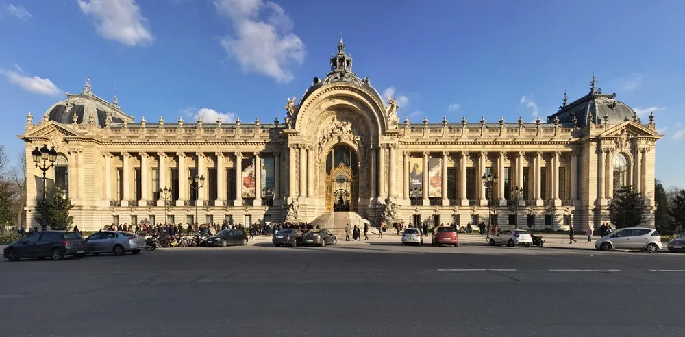 Paris'teki görkemli Petit Palais müzesinin altın sarısı taş cephesi, güneşli bir havada, merkezi kubbesi, detaylı oymaları ve sütunlarıyla ön plana çıkarken, önündeki cadde ve kalabalık ziyaretçilerle birlikte geniş bir panoramik görüntü sunuyor.