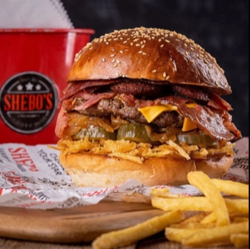 Susamlı hamburger ekmeği arasında kalın köftesi, erimiş çedar peyniri, karamelize soğan, turşular ve bol miktarda dana pastırma ile servis edilen, yanında çıtır patates kızartması ve Shebo’s logolu kırmızı bir kova bulunan bu iştah açıcı hamburger, İzmir'in en iyi restoranları arasında anılan Shebo's Sandwich Burger'ın lezzet duraklarından birini yansıtıyor.