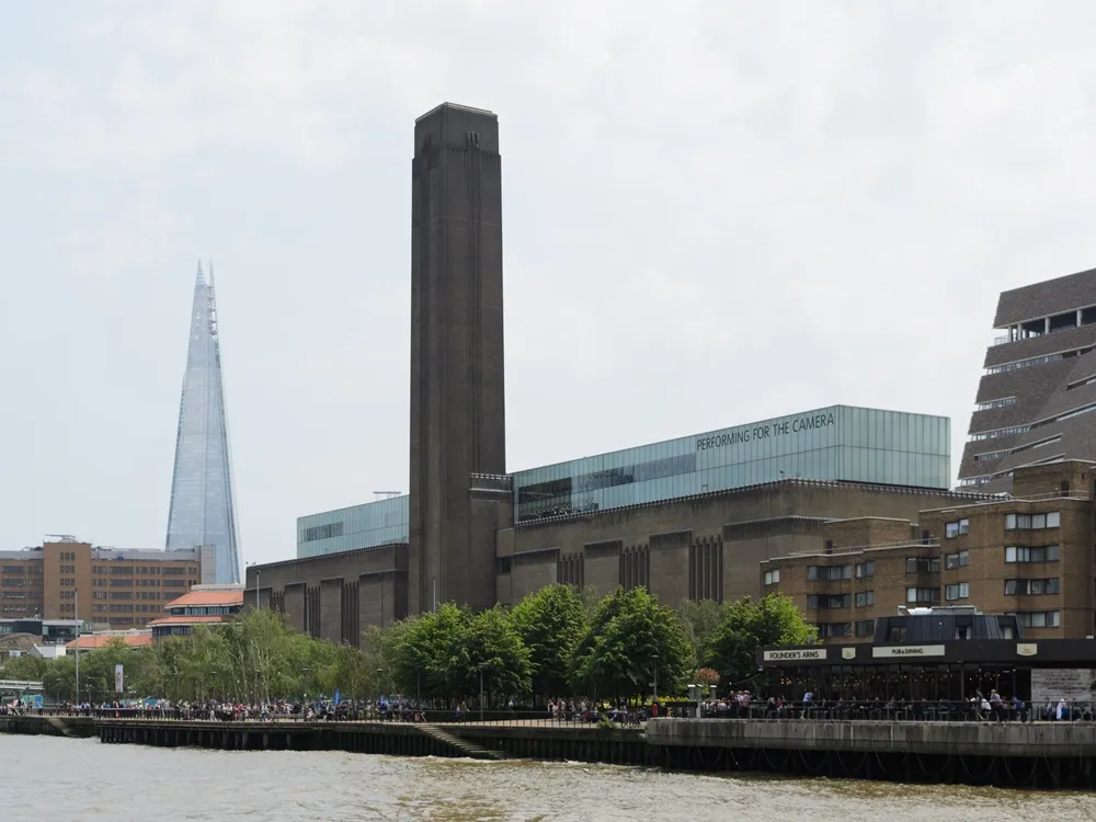 Thames Nehri'nin üzerinden çekilmiş geniş açılı bir fotoğrafta, ikonik Tate Modern binasının uzun bacası ve üzerinde PERFORMING FOR THE CAMERA yazısı bulunan cam yapısı, solunda sivri uçlu Shard gökdeleni ile birlikte Londra'nın hareketli nehir kıyısı boyunca uzanan kalabalık yürüyüş yolunun ve Founder's Arms gibi çeşitli yapıların üzerinde yükseliyor.