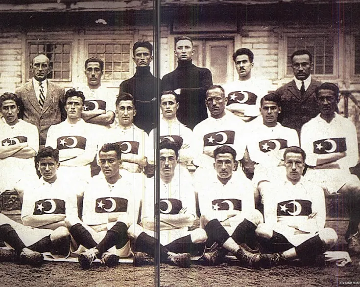Siyah beyaz, eski bir fotoğrafta, 1924 yılına ait Türkiye'nin ilk Milli Takımı oyuncuları, göğüslerinde ay yıldız arması bulunan beyaz formalarıyla ciddi ifadelerle poz vermiş, arka planda ahşap bir yapı yer alıyor.