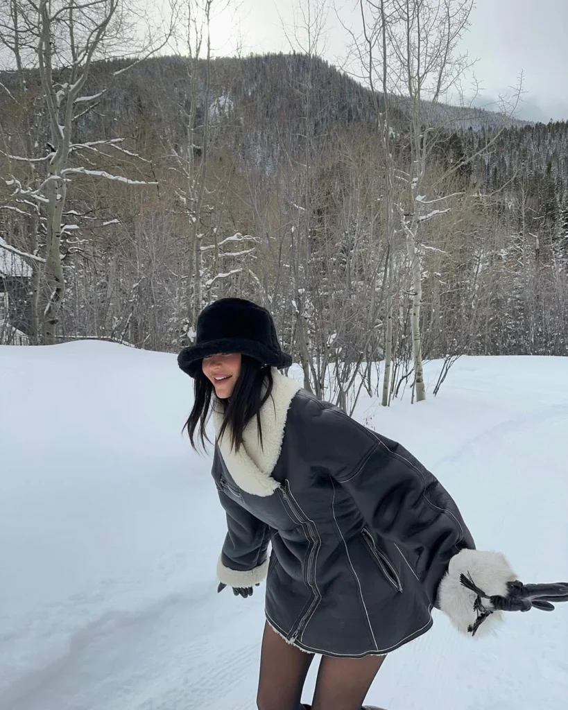 Karlı doğada hareket halinde poz veren Kylie Jenner; siyah shearling detaylı mont ve mini elbiseyle cool ve modern après-ski görünümü.