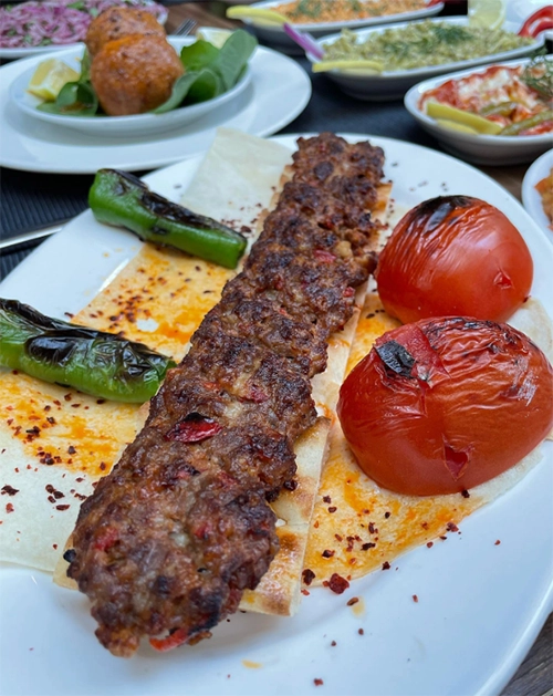 Yanında közlenmiş domates ve biberlerle, lavaş üzerinde sunulan, kömür ateşinde özenle pişirilmiş iştah açıcı Adana kebap, arka planda çeşitli mezelerle İzmir'in en iyi kebap restoranlarından birindeki lezzetli bir ocakbaşı deneyimini yansıtıyor.