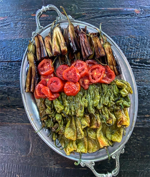 Koyu renk ahşap bir masa üzerinde duran gümüş oval tepside, nar gibi kızarmış patlıcan, dilimlenmiş domates ve yeşil biberlerin iştah açıcı düzenlemesi, İzmir'in zengin gastronomi sahnesinden lezzetli bir garnitür veya meze sunumunu yansıtır.