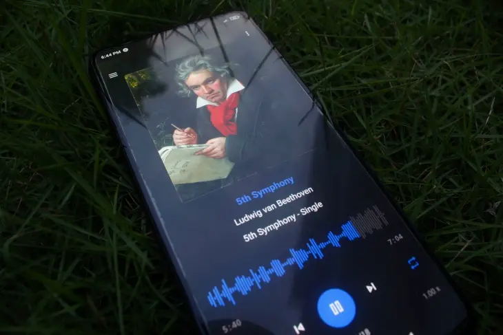 Yeşil çimlerin üzerinde duran bir akıllı telefon ekranında, Ludwig van Beethoven'ın 5. Senfonisi çalarken, bestecinin elinde notalarla kalem tuttuğu bir portresi belirgin biçimde yer alıyor, bu da Beethoven'ın eserlerini çağdaş bir şekilde deneyimlemeyi yansıtıyor.