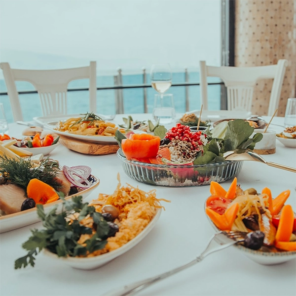 İzmir'in en iyi restoranlarından birinde, denizin nefes kesen manzarasına karşı kurulmuş beyaz örtülü bir masada, nar taneleri ve yeşilliklerle süslenmiş salata, balık marinleri, humus ve makarnanın da dahil olduğu Ege mutfağından çeşitli taze ve iştah açıcı mezelerin ve bir kadeh beyaz şarabın bulunduğu keyifli bir ziyafet masası gösterilmektedir.