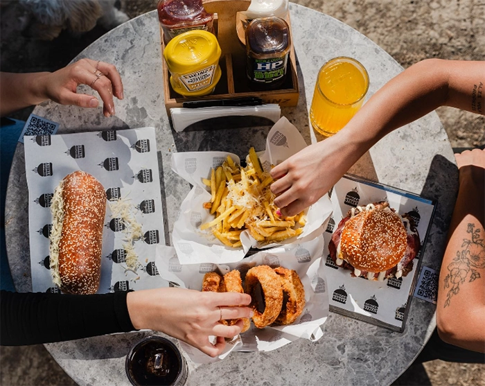 Üstten çekilmiş bir fotoğrafta, İzmir'in en iyi restoranları arasında yer alan Burger Street'ten gelen bir burger, bol peynirli patates kızartması, soğan halkaları ve uzun bir sandviçin etrafında toplanmış üç kişi, masadaki içecekler ve soslarla birlikte keyifli bir öğün deneyimi yaşarken görülüyor.