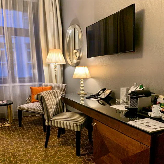 Ahşap ve parlak yüzeyli bir çalışma masası, iki şık sandalye, yumuşak ışık veren lambalar ve duvardaki televizyonla oval aynanın olduğu bu konforlu otel odası, Prag seyahati için ideal bir konaklama seçeneği sunuyor.