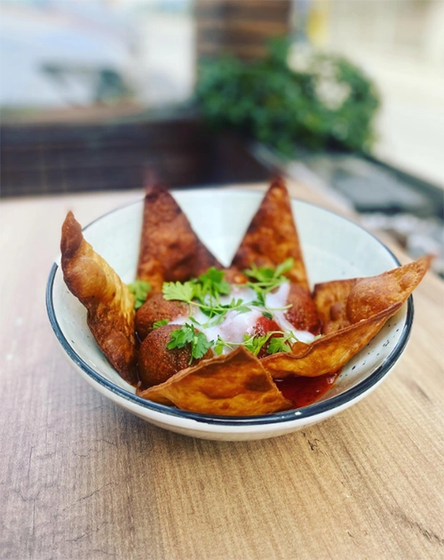 İzmir'deki en iyi restoranlardan biri olan Cute Gastro Pub'ın menüsünden, ahşap bir zeminde duran, kenarları çıtır çıtır kızarmış hamur parçalarıyla çevrili derin bir kasenin içinde, üzerine beyaz sos ve taze maydanoz serpilmiş, iştah açıcı falafel topları içeren bir vegan yemek.