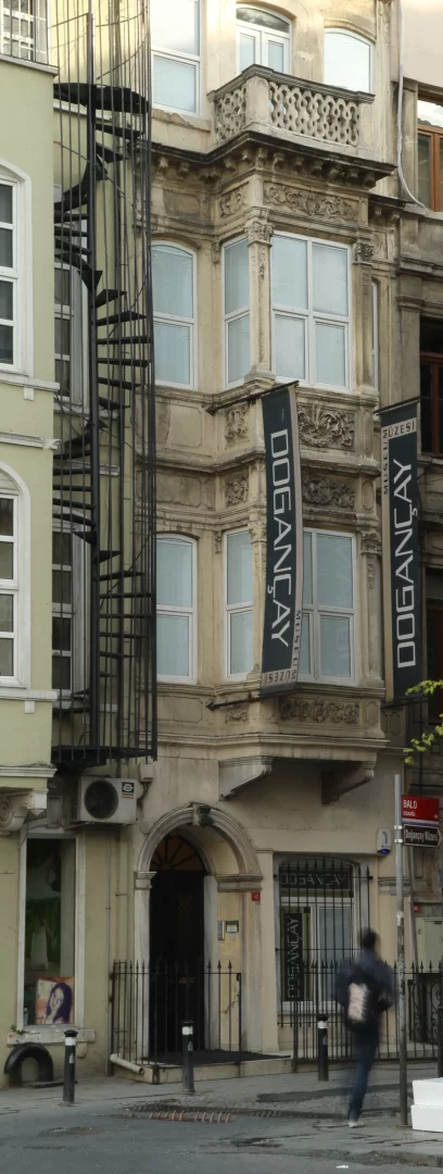 Beyoğlu'ndaki Doğançay Müzesi'nin, cephesi detaylı süslemelerle bezeli, çok sayıda pencereli, alt katında cumba pencereleri ve üstünde oymalı korkuluklu balkonu olan tarihi binası, yanında uzanan siyah metal spiral merdivenle birlikte, kaldırımda yürüyen bulanık bir kişiyle birlikte gün ışığında görünüyor.