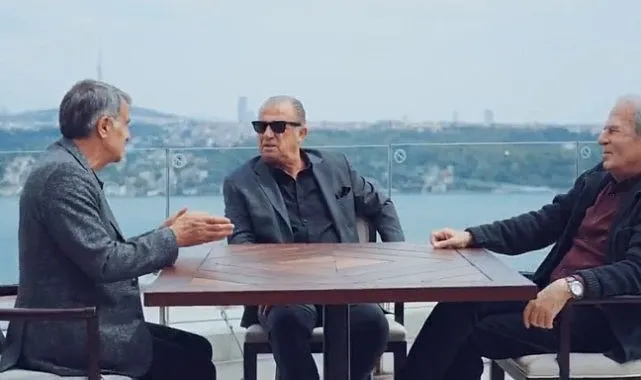 Türk futbolunun üç önemli teknik direktörü, solda oturan Şenol Güneş, güneş gözlüklü Fatih Terim ve sağda Mustafa Denizli, İstanbul Boğazı manzaralı açık bir terasta ahşap bir masanın etrafında toplanmış, derin bir sohbetin içindeler.