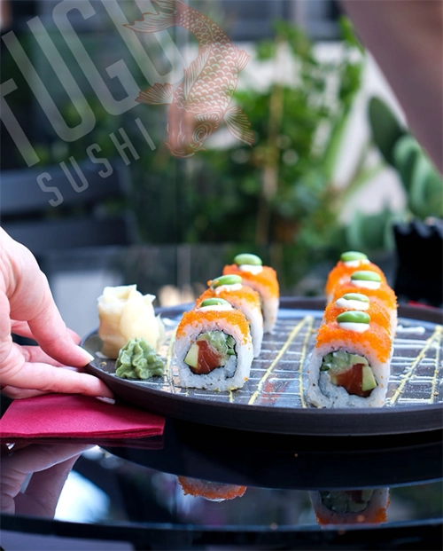 İzmir'in en iyi sushi restoranlarından My Fugu Sushi'de sunulan, parlak turuncu tobiko, avokado, somon ve üzerine yerleştirilmiş edamame tanesi ile göz alıcı bir şekilde hazırlanan sushi roll tabağı, taze zencefil ve wasabi eşliğinde, yeşil bitki örtüsüyle çevrili şık bir masada bir el tarafından özenle yerleştirilirken şehrin gastronomi zenginliğini yansıtıyor.