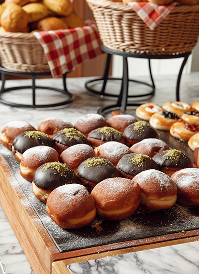 Büyük bir brunch büfesinin tatlı bölümünde sergilenen, üzeri pudra şekeriyle süslenmiş, çikolata soslu ve antep fıstıklı gibi farklı çeşitlerdeki bol miktarda lezzetli çörek ve donutların cazip görünümü.