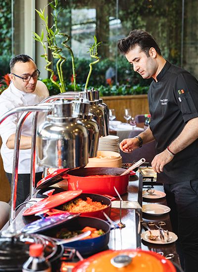 Hyatt'ın zengin brunch büfesinde, siyah üniformalı bir şef çeşitli sıcak yemekleri özenle servis ederken, beyaz üniformalı şefin dikkatli denetimi altında misafirler için unutulmaz bir lezzet deneyimi sunuluyor.