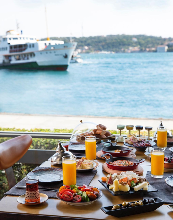 Boğaz manzarasında, üzerinde zeytin, peynir çeşitleri, domatesli salata, yumurta tavaları, portakal suyu bardakları ve çay bulunan zengin bir kahvaltı masası ile arkada beyaz bir vapurun geçtiği masmavi sular görülüyor.