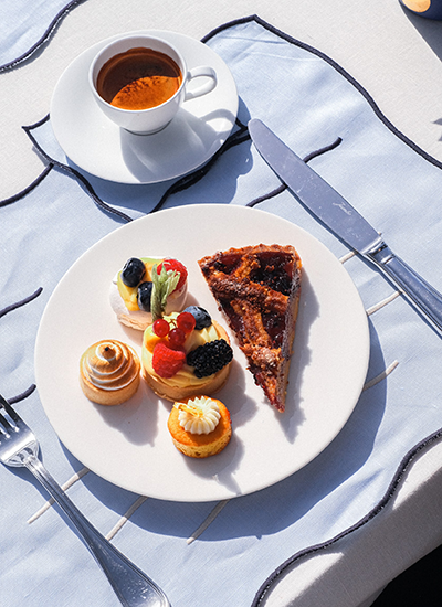 Parlak güneş ışığı altında, beyaz bir tabakta frambuaz, böğürtlen ve kivi dilimleriyle süslenmiş üç küçük meyveli tart, spiral şekilli bir beze, üzeri kremalı diğer bir mini pasta ve koyu renkli bir tart diliminin iştah açıcı bir şekilde sunulduğu, yanında metal çatal bıçak takımı, dalgalı desenli açık mavi masa örtüsü ve arka planda bir fincanın seçildiği zengin tatlı tabağı.