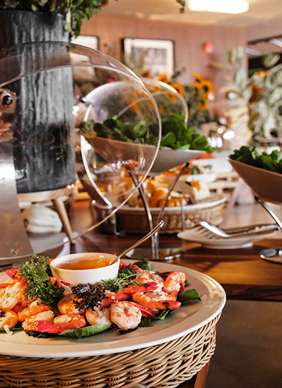 Lacivert'in zengin brunch menüsünden bir kesit sunan bu karede; ön planda taze karidesler, çeşitli yeşillikler ve özel sos ile hazırlanan iştah açıcı bir tabak, arka planda ise şık cam kaplı katlı büfe düzeninde sunulan zengin salata ve başlangıç seçenekleri zarif bir sunumla dikkat çekiyor.