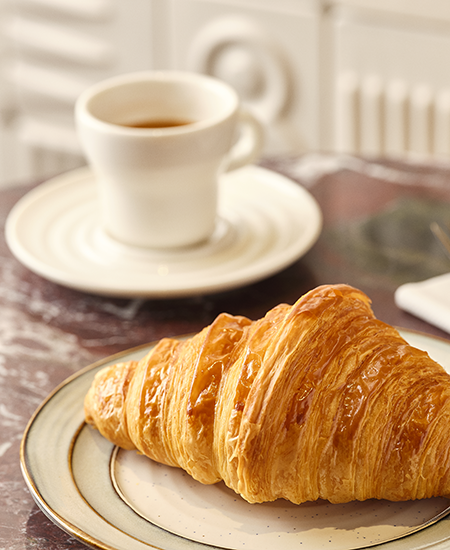Desenli kenarlı beyaz bir tabağın üzerinde yer alan, katmanlı yapısıyla dikkat çeken, altın rengi bir kruvasan ve arka planda mermer yüzeyde duran, koyu kahve içeren beyaz bir espresso fincanının yakın çekim görüntüsü.