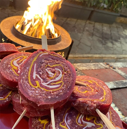 Yakın çekimde, ahşap şişlere takılı, içinde sarı ve beyaz dolgularla spiral desen oluşturan, ızgaraya hazır çiğ et sarmaları ön planda dururken, bulanık arka planda yanan bir ateş çukuru İzmir'in en iyi et restoranları arasında yer alan bir mekandaki özenle hazırlanmış et lezzetlerini gösteriyor.