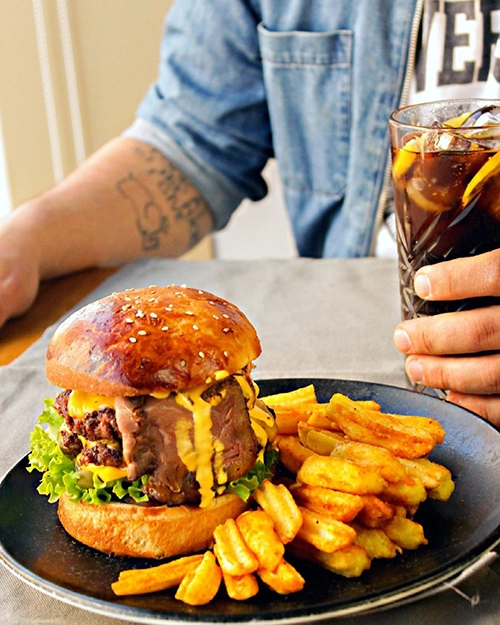 İzmir'in en iyi hamburgercileri arasında yer alan Mengoli Burgers'ın kendi mutfaklarında pişirdiği susamlı ekmeği, eritilmiş bol çedar peyniriyle lezzetlendirilmiş dana bonfile dilimleri ve sulu köftesiyle dolu gurme hamburgeri, yanında taptaze kızarmış patatesleri ve buzlu içeceğiyle iştah açıcı bir sunumda.