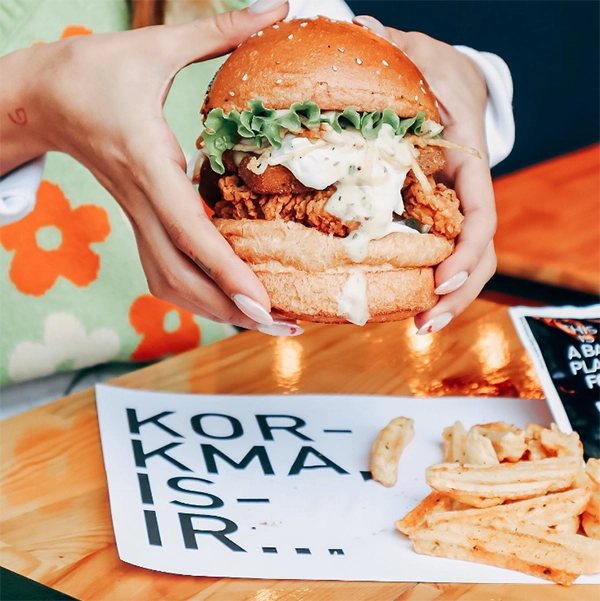 Bir kişinin ellerinde tuttuğu, çıtır kızarmış tavuk, taze marul ve bol kremalı sosla dolu, pofidik ekmekli, iştah açıcı büyük bir hamburger, yanında baharatlı patates kızartmaları ve üzerinde KORKMA! İZMİR... yazan bir kağıtla sunularak, İzmir'in en iyi restoranları arasında öne çıkan zengin hamburger çeşitliliğini ve canlı gastronomi sahnesini gözler önüne seriyor.