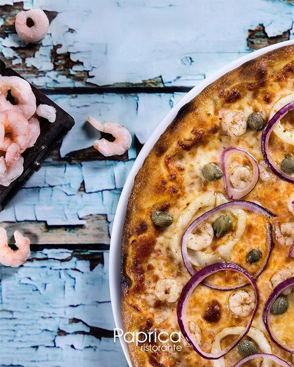 İzmir'in en iyi restoranları arasında yer alan Paprica Ristorante'den, taze karides, kırmızı soğan ve kapariyle bolca donatılmış, erimiş peynirle kaplı, altın sarısı kenarlı bir pizzanın iştah açıcı yakın çekim görüntüsü, yanında tahta bir kesme tahtası ve yıpranmış mavi ahşap zeminde pembe karideslerle tamamlanıyor.