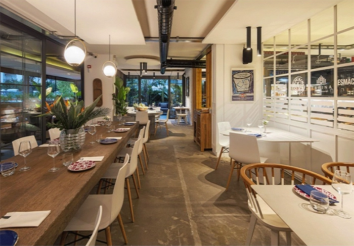 İzmir'in en iyi restoranlarından biri olan Peperino Pizza Italiana'nın modern iç mekanı, uzun ahşap yemek masası, cam duvarlardan yansıyan yeşillikler ve zarif beyaz sandalyelerle misafirlerini ağırlamaya hazır şık bir atmosfer sunuyor.