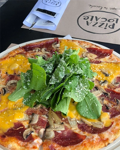 İzmir'in en iyi pizzacılarından biri olan Pizza Locale'den, markalı kağıt altlık ve peçetelik önünde, ince hamurunda dilimlenmiş mantarlar, şarküteri ürünleri ve bol erimiş sarı peynirle hazırlanmış, ortası rendelenmiş parmesan peyniri serpilmiş taze roka yığınıyla dolu, çok çeşitli ve iştah açıcı bir pizza yakın plan görünüyor.