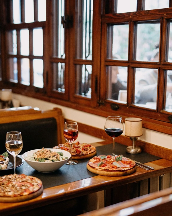 İzmir'in en iyi restoranları listesinde yer alan popüler bir pizzacının iç mekanında, ahşap çerçeveli büyük pencerelerin önündeki bir masada mantarlı, salamlı ve domatesli gibi çeşitli lezzetli pizzalar, taze yeşilliklerle dolu bir salata kasesi ve farklı renkteki şarap kadehleri zarif bir lamba eşliğinde servis edilmiş, dışarıdaki hareketli şehir yaşamı da pencerelerden hafifçe yansıyor.