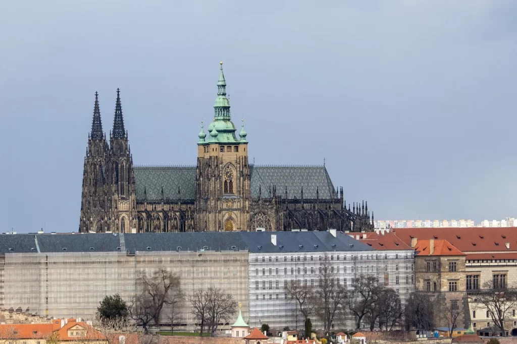 Bulutlu gökyüzü altında, Prag Kalesi’nin görkemli bir parçası olan, koyu renkli sivri kuleleri ve yeşil kubbeleriyle Aziz Vitus Katedrali'nin tarihi ihtişamı, ön plandaki iskeleli binaların ve çıplak ağaçların arasından bu mistik şehrin simgesel güzelliğini yansıtıyor.