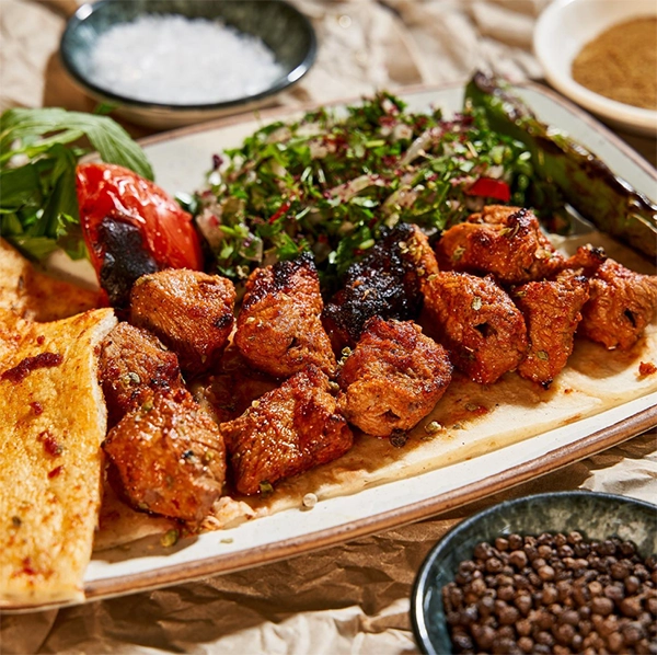 İzmir'in en iyi restoranlarından birinde sunulan, özel baharatlarla terbiye edilmiş közlenmiş kuzu şişler, yanında bol yeşillikli sumaklı salata, ızgara domates ve biberle iştah açıcı bir sunumla servis ediliyor.