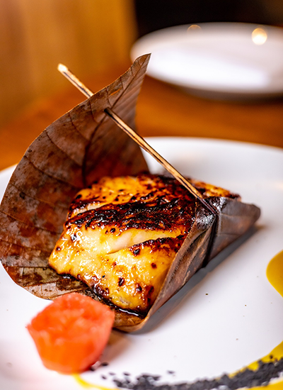 ROKA BRUNCH menüsünden, özel bir yaprağa sarılıp odun şişle tutturulmuş, üzerinde hafif kömürleşmiş ızgara izleri olan, beyaz tabakta servis edilen, yanında kırmızımsı bir garnitür ve sarı sos ile sunulan, lezzetli görünüşlü bir balık yemeği detaylıca gösteriliyor.