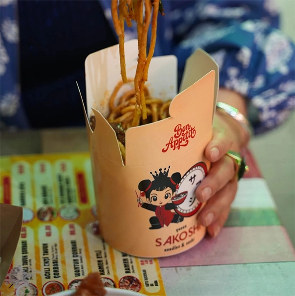 Bir kişi, üzerinde Sakoshi markasının sevimli bir kız figürü ve logosu bulunan karton kutudan chopstick'lerle buharlı, lezzetli noodle'ları kaldırırken, arka planda Uzak Doğu mutfağı menüsü görünen bu sahne, İzmir'in en iyi restoranlarından Sakoshi'nin otantik lezzetlerini vurguluyor.