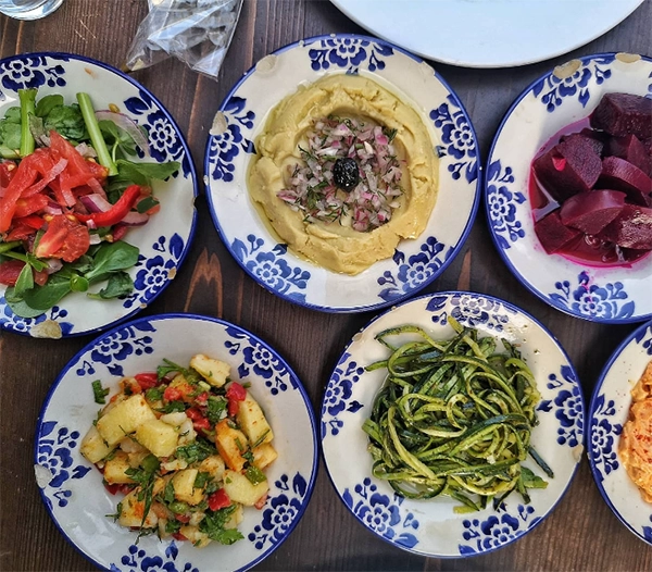 Koyu ahşap bir masanın üzerinde, İzmir'in en iyi restoranlarında sunulan ve yöresel lezzetleri yansıtan, mavi-beyaz desenli tabaklarda sergilenen zengin bir meze sofrası; taze yeşillikli salata, zeytinyağlı fava, parlak mor pancar turşusu, patates salatası, kabak spagetti ve kırmızımsı bir dip ile damakları şenlendiriyor.