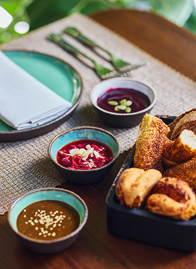 Ahşap bir masanın üzerinde sunulan zengin bir brunch masası detayı, içerisinde çeşitli altın rengi ekmekler ve çıtır hamur işlerinin bulunduğu siyah bir sepeti gösteriyor; yanında, yeşil kabak çekirdekleriyle süslenmiş mor renkli bir meyve sosu, kıyılmış bademlerle bezenmiş parlak kırmızı bir reçel ve beyaz susam taneleriyle zenginleştirilmiş kahverengi bir tahin veya fındık ezmesi olmak üzere üç farklı lezzetli dip sos küçük seramik kaselerde özenle yer alıyor.