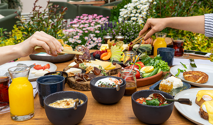 Çiçekler ve yeşillikler içindeki dış mekan bir masada, çeşitli peynirler, şarküteri ürünleri, taze meyveler, sebzeler, simit, bal peteği, Eggs Benedict ve içeceklerle dolu zengin bir serpme kahvaltıda iki kişi maşalarla yemek seçiyor.