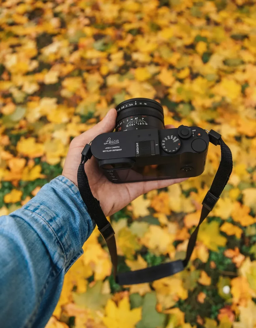 Kot ceketli bir el, arka planda sarı ve turuncu sonbahar yapraklarıyla kaplı zeminin üzerinde, siyah bir Leica fotoğraf makinesi tutuyor ve sonbahar fotoğrafçılığı için hazır bir anı yansıtıyor.