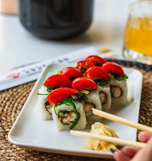 Beyaz pirinç, kızarmış dolgu, yeşil deniz yosunu ve yarıya bölünmüş parlak kırmızı çeri domateslerle hazırlanan lezzetli sushi rulolarının beyaz dikdörtgen bir tabakta özenle sunulduğu, sağda bir elin turşulu zencefili tutan yemek çubuklarını gösterdiği bu kare, İzmir'deki en iyi restoranlardan birinde Uzak Doğu mutfağının özel sunumlarını yansıtmaktadır.