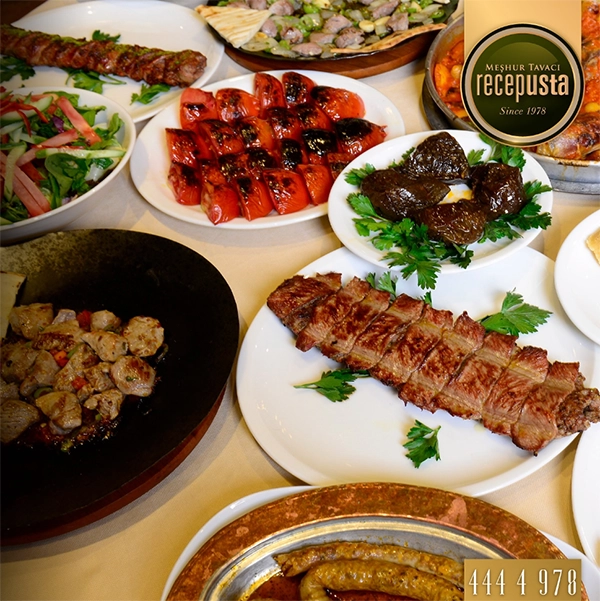 Meşhur Tavacı Recep Usta logosunun da yer aldığı bu gösterişli masada, ızgara kaburga, Adana kebap, közlenmiş domatesler, pirinçli yaprak sarmaları ve tava yemeği gibi iştah açıcı et ve meze çeşitleriyle dolu geleneksel bir ziyafet sofrası, İzmir'in en iyi restoranları arasındaki kalitesini yansıtıyor.