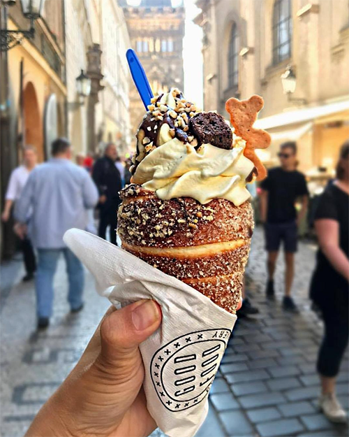 Prag'ın tarihi bir sokağında bir elin tuttuğu, içi dondurma, çikolata sosu, kıyılmış fındık, brownie ve kurabiye ile zenginleştirilmiş meşhur Trdelník tatlısı, şehrin mistik güzelliğini ve canlılığını yansıtan kalabalık bir arka planla Prag seyahat rehberi için iştah açıcı bir sahne oluşturuyor.
