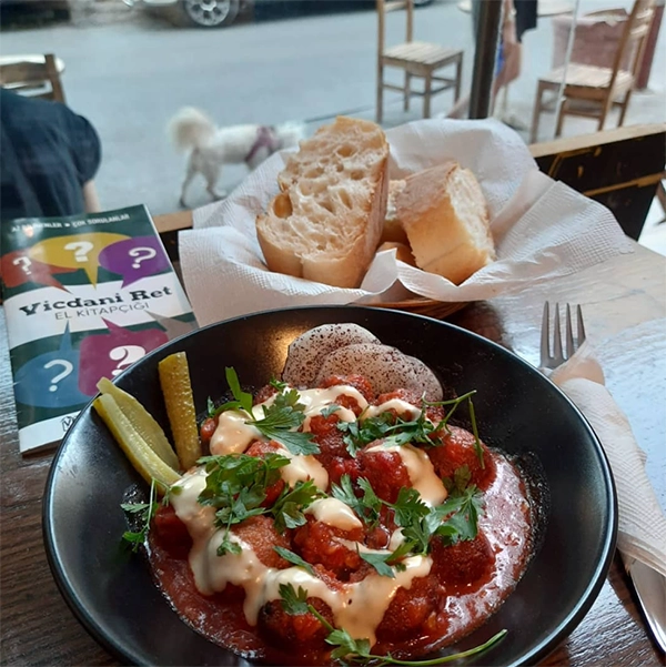 İzmir'in en iyi restoranları arasında yer alan ve vegan lezzetleriyle öne çıkan Tyke Vegan Kafe'de, domates soslu fellah köftesi veya falafelin üzerinde beyaz sos ve maydanozla sunulduğu, yanında taze ekmek ve turşu bulunan, ayrıca Vicdani Ret El Kitapçığı'nın da masada olduğu bir yemek anı.