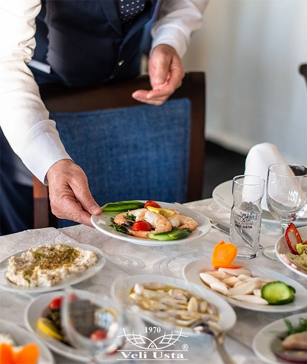 Bir garsonun beyaz örtülü masaya taze karides, çeri domates ve salatalık dilimleriyle süslenmiş bir meze tabağını dikkatlice bıraktığı Veli Usta Körfez Restoran'da, İzmir'in en iyi balık restoranları listesinde adı geçen deniz ürünleri ve çeşitli mezelerle donatılmış zengin bir sofra manzarası.