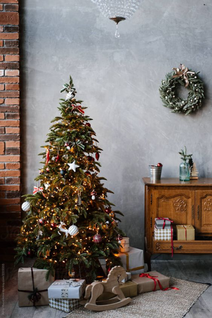 Minimal ve doğal yılbaşı dekorasyonu; ışıklı Noel ağacı, hediye paketleri ve tuğla duvar detayları.